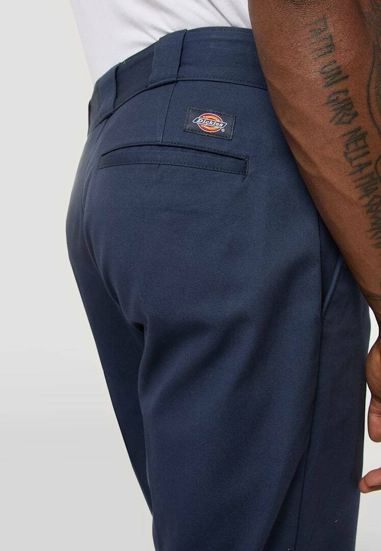 Close-up of person wearing navy blue Dickies pants, showing back pocket and branded label, with tattooed arm visible.