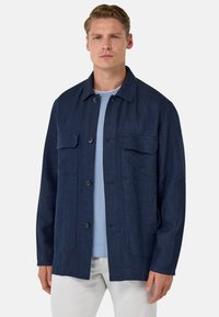 Marineblauwe jas van textiel met structuur. Het heeft een knoopsluiting aan de voorkant, twee borstzakken en een ontspannen pasvorm met een kraag.