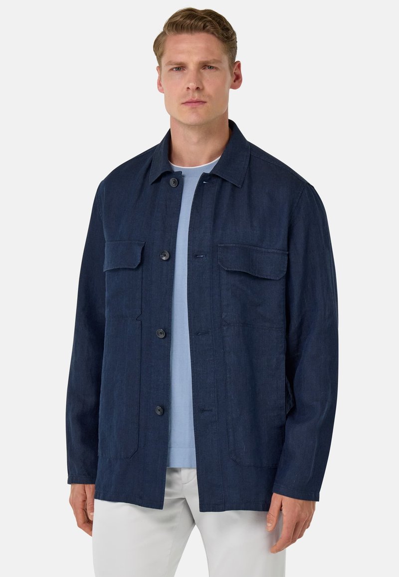 Marineblauwe jas van textiel met structuur. Het heeft een knoopsluiting aan de voorkant, twee borstzakken en een ontspannen pasvorm met een kraag.
