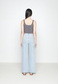 Žena s dlouhými černými vlasy, oblečená v šedém tílku a světle modrých širokých kalhotách, stojí čelem k bílé zdi.