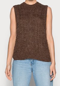 Brauner ärmelloser Zopfmuster-Pullover mit rundem Halsausschnitt und strukturierter Optik, kombiniert mit hellblauen Jeans.