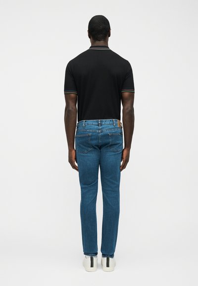 Άνδρας με την πλάτη, που φοράει μαύρο κοντομάνικο πουκάμισο με ριγέ μανσέτες, μπλε τζιν και λευκά αθλητικά παπούτσια με μαύρες ρίγες.