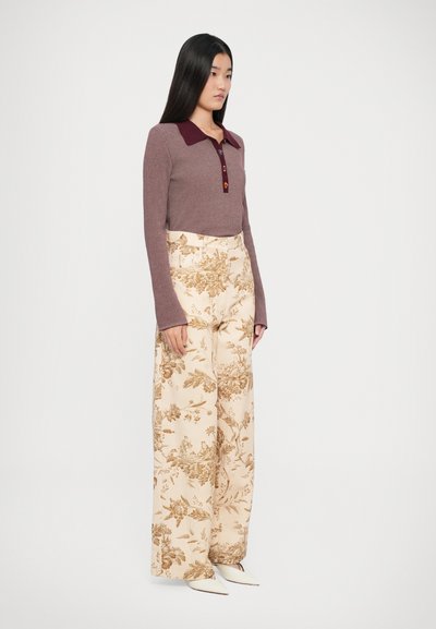 Polo long à manches longues côtelé bordeaux avec patte de boutonnage, associé à un pantalon large crème arborant un motif floral marron. Chaussures blanches à bouts pointus.