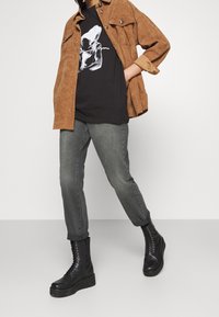 Chaqueta de pana marrón, camiseta gráfica negra, jeans grises ajustados, botas negras con cordones y suelas de plataforma. Conjunto casual en capas.