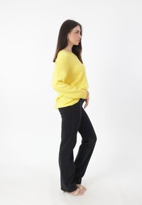 Gelber gestrickter Pullover mit langen Ärmeln, locker geschnitten. Kombiniert mit hochgeschnittenen schwarzen Jeans und hellen Sandalen.