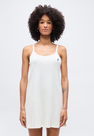 Femme aux cheveux bouclés portant une robe blanche texturée sans manches avec un petit logo sur la poitrine, debout devant un fond clair uni.