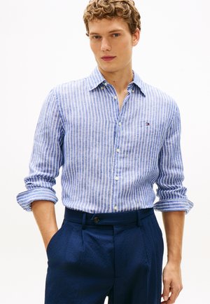 Młody mężczyzna z podwiniętymi rękawami, ubrany w niebiesko-białą pasiastą koszulę zapinaną na guziki i granatowe spodnie, ręce w kieszeniach, neutralne tło.