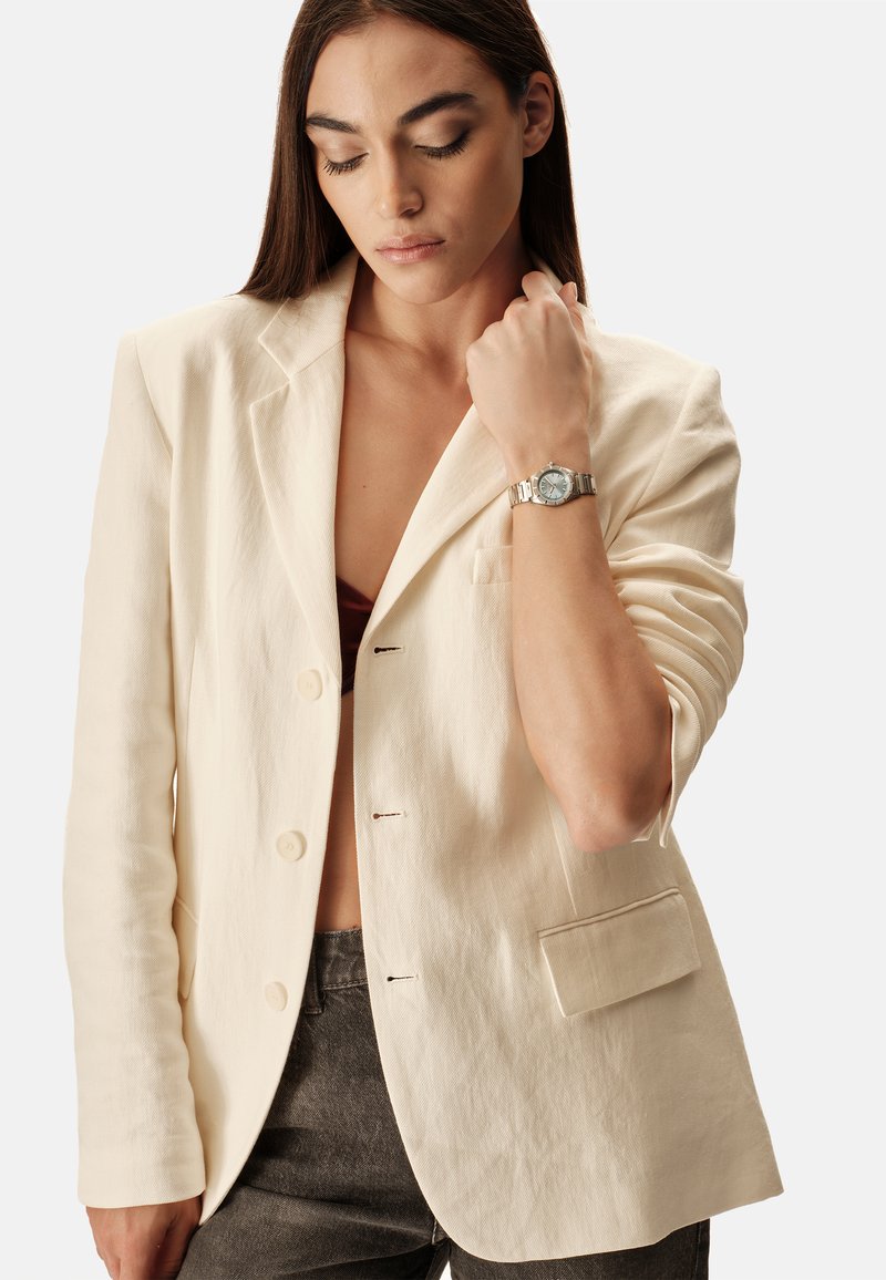 Linnen blazer in roomkleur met een getailleerd ontwerp, twee knopen en een enkele borstzak. Gecombineerd met een bordeauxrode top en grijze spijkerbroek.