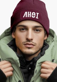 Maroon knit beanie with "AHOI" and anchor design, worn with a green puffer jacket. Close-up view of face, showcasing texture and details.