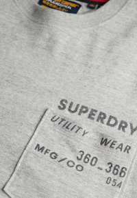 Grå sweatshirt med struktureret materiale, ribbet halsudskæring og en frontlomme med trykte 'SUPERDRY', 'UTILITY WEAR' og produktionsdetaljer.