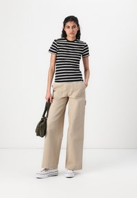 T-shirt a maniche corte a righe bianche e nere abbinato a pantaloni beige a gamba larga e sneakers bianche. Il modello tiene in mano una piccola borsa verde.