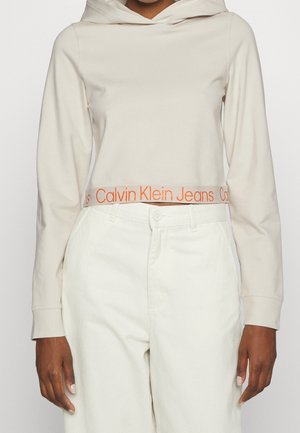 Henkilö, jolla on päällään vaaleabeige lyhyt huppari, jossa on oranssi Calvin Klein Jeans -logo helmassa, ja korkeavyötäröiset luonnonvalkoiset housut.
