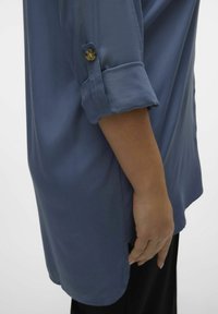 Chemise bleue avec des manches retroussées, des détails de boutons et une texture douce. Coupée longue avec un ourlet arrondi, assortie à un pantalon noir.