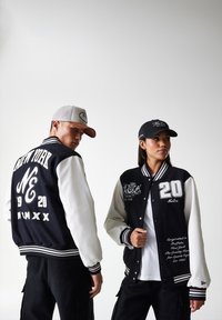 Black and white varsity jackets feature embroidery and stripes, with white sleeves. Models wear black and gray caps with logos.