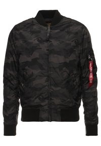 Veste bomber camouflage noire et grise avec fermeture éclair avant, poignets et ourlet côtelés, et étiquette rouge "Remove Before Flight" sur la fermeture éclair de la poche de la manche gauche.