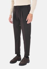 Pantaloni marroni plissettati con tasche laterali, chiusura a bottone e dettagli con cordino. Abbinati a scarpe in suede marroni scuri.