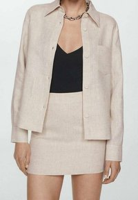 Camicia button-up in lino beige con colletto, abbinata a una gonna fitted coordinata. Top nero sotto, con un dettaglio tasca discreto sulla camicia.