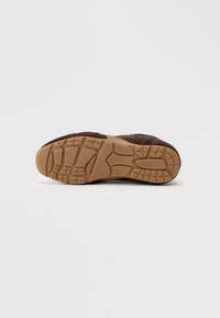 Sapatilha de camurça castanha com uma sola de borracha texturizada, apresentando sulcos profundos e uma biqueira arredondada. Acabamento mate geral com detalhes de design subtis.