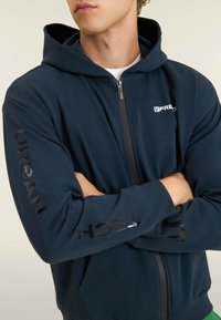 Giovane che indossa una felpa con zip di colore blu navy con la scritta "Urban Tech" sulla manica e un piccolo logo sul petto, braccia incrociate.