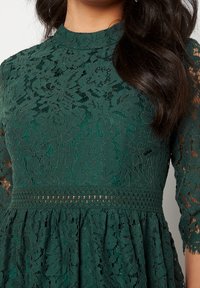 Robe en dentelle verte avec un col montant et des manches trois-quarts. Motif floral, tissu texturé et taille soulignée par des détails froncés.