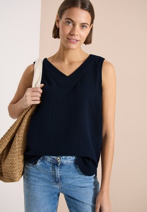 Jeune femme portant un haut noir sans manches à col en V et un jean bleu, tenant un sac fourre-tout tissé sur son épaule, sur un fond neutre.
