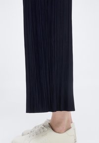 Κάτω άκρα που φορούν μαύρο πλισέ παντελόνι και λευκά αθλητικά παπούτσια με κορδόνια σε λευκό φόντο.