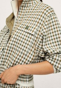 Zielono-biała kurtka w wzór houndstooth z wysokim kołnierzem, z zamkiem błyskawicznym, przednią kieszenią i teksturowanym materiałem.