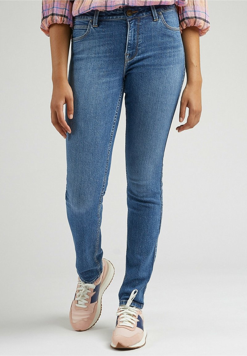 Jean skinny bleu en denim, avec une taille mi-haute et un design à cinq poches, associé à des baskets rose clair avec des accents blancs.
