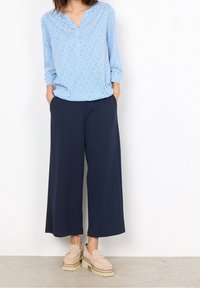 Blusa azzurro chiaro con motivo geometrico bianco, abbinata a pantaloni a gamba larga blu scuro. Mocassini beige con suola spessa completano l'outfit.