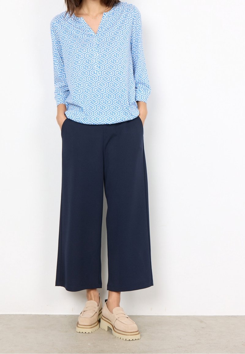 Blusa azzurro chiaro con motivo geometrico bianco, abbinata a pantaloni a gamba larga blu scuro. Mocassini beige con suola spessa completano l'outfit.