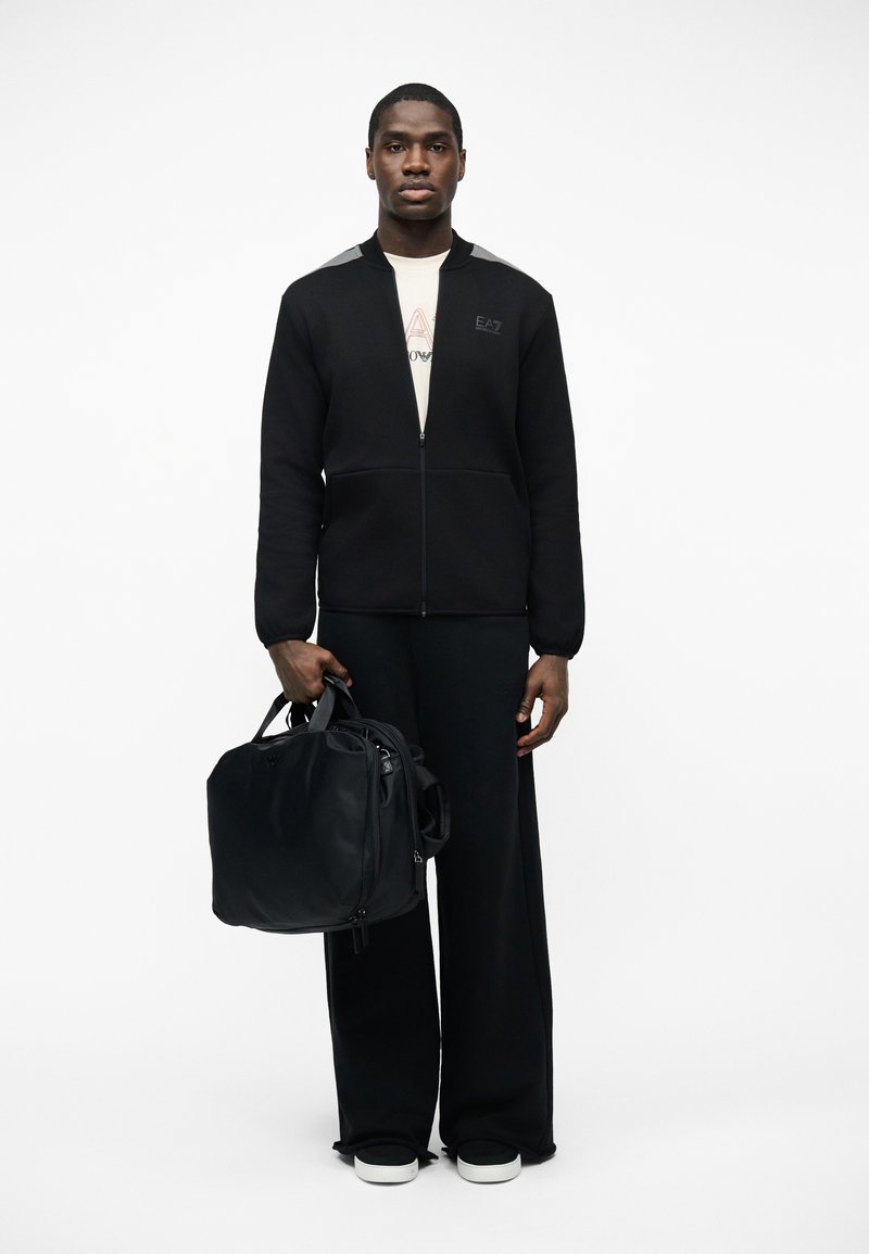 Uomo in piedi che indossa una giacca nera con zip, pantaloni neri larghi, scarpe da ginnastica bianche, che tiene una grande borsa nera nella mano sinistra.