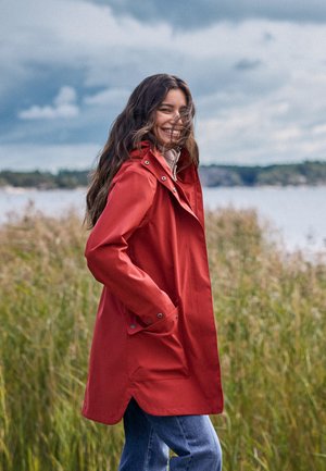 Uśmiechnięta kobieta w czerwonym płaszczu przeciwdeszczowym i dżinsach stoi na trawiastej przestrzeni blisko wody, pod zachmurzonym niebem, z rękami w kieszeniach płaszcza.