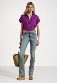 Lilla kortermet skjorte, denim flared jeans, vevd belte, stråveske og beige kilehæler, presentert mot en hvit bakgrunn.