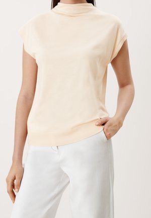 Krótki top z krótkim rękawem w jasnym brzoskwiniowym kolorze, z wysokim kołnierzem i ściągaczem na dole, wykonany z gładkiej, miękkiej tkaniny. Połączony z szerokimi białymi spodniami.