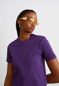 Camiseta púrpura de manga corta con cuello redondo y logo bordado. El modelo lleva gafas de sol transparentes amarillas con acentos beige.