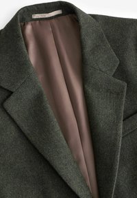 Grøn uldblandet blazer med reverskrave og en glat, lys brun satinforing synlig indeni. Enkel, skræddersyet design uden pynt.