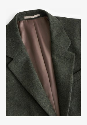 Vihreä villasekoitteinen bleiseri, jossa on viistetty kaulus ja sisällä näkyvää sileää vaaleanruskeaa satiinia. Yksinkertainen, räätälöity muotoilu ilman koristeita.