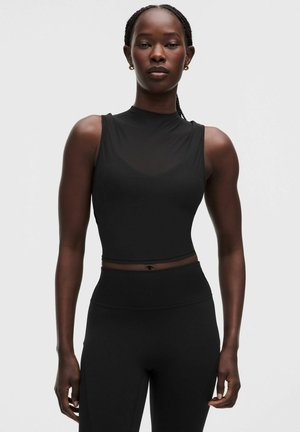 Kobieta z warkoczami na włosach nosi bezrękawnikowy czarny crop top z wysokim kołnierzem i czarne legginsy z wysokim stanem, stojąc na jednolitym tle.