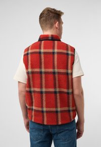Rote Fleece-Weste mit karriertem Muster in Beige und Marineblau, mit Kragen und ohne Ärmel. Getragen über einem cremefarbenen Hemd.