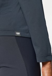 Námornícka športová dlhá košeľa s plochými švami, spárovaná s úzkymi nohavicami v rovnakej námorníckej farbe. Obsahuje malé logo ciele Athletics.