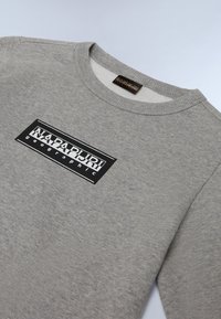 Sudadera gris con cuello redondo acanalado. Presenta un prominente parche de logo en blanco y negro en la parte delantera. Tejido suave y texturizado.