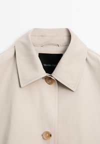 Manteau beige à col avec deux boutons marron visibles et une étiquette noire à l'intérieur de l'encolure portant l'inscription "Massimo Dutti".
