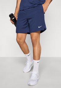 Αθλητικά σορτς σε ναυτικό μπλε χρώμα από ελαφρύ ύφασμα, με πλευρικές σκισμές και διακριτικό λογότυπο Nike, συνδυασμένα με λευκά αθλητικά παπούτσια και καλτσάκια.