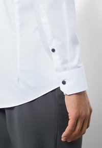 Weißes Hemd aus glattem, leichtem Stoff, mit kontrastierenden dunklen Knöpfen und einem Logo an der Manschette, kombiniert mit einer dunklen Hose.