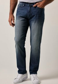 Jeans de mezclilla azul oscuro con un lavado degradado, diseño de cinco bolsillos y cierre de botón. Llevados con zapatillas blancas.