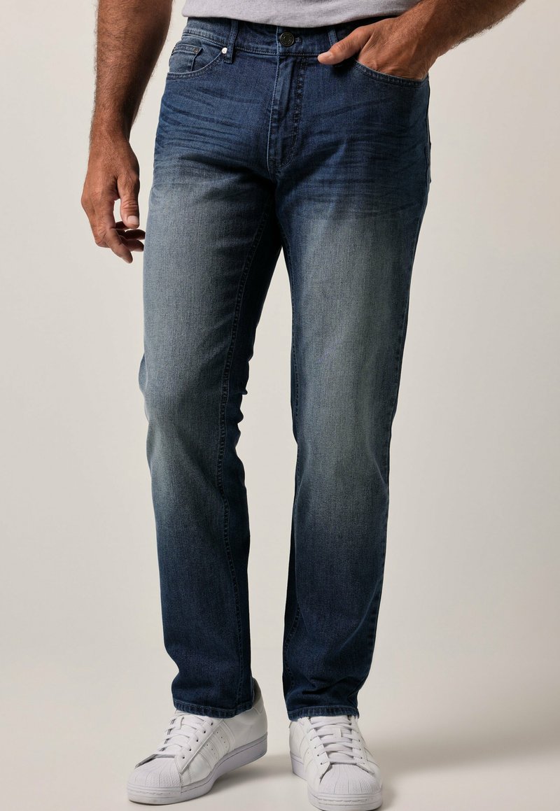 Jeans de mezclilla azul oscuro con un lavado degradado, diseño de cinco bolsillos y cierre de botón. Llevados con zapatillas blancas.