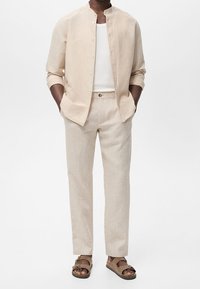 Homme portant une chemise beige boutonnée par-dessus un débardeur blanc, un pantalon beige assorti et des sandales marron, debout les mains dans les poches.