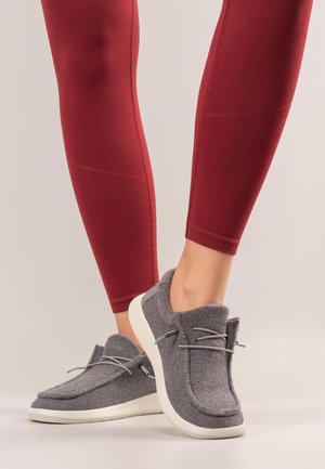 Beine in roten Leggings, die graue Slip-on-Freizeitschuhe mit weißen Sohlen tragen, stehend auf einem hellen neutralen Hintergrund.