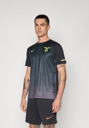 Mann in schwarzem Sportoutfit mit neon-gelben Logos und Tätowierungen auf beiden Armen, der vor einem schlichten hellen Hintergrund steht.