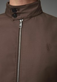 Veste marron avec un col haut, deux boutons noirs et une fermeture éclair argentée à l'avant ; texture lisse avec un détail de logo en relief.